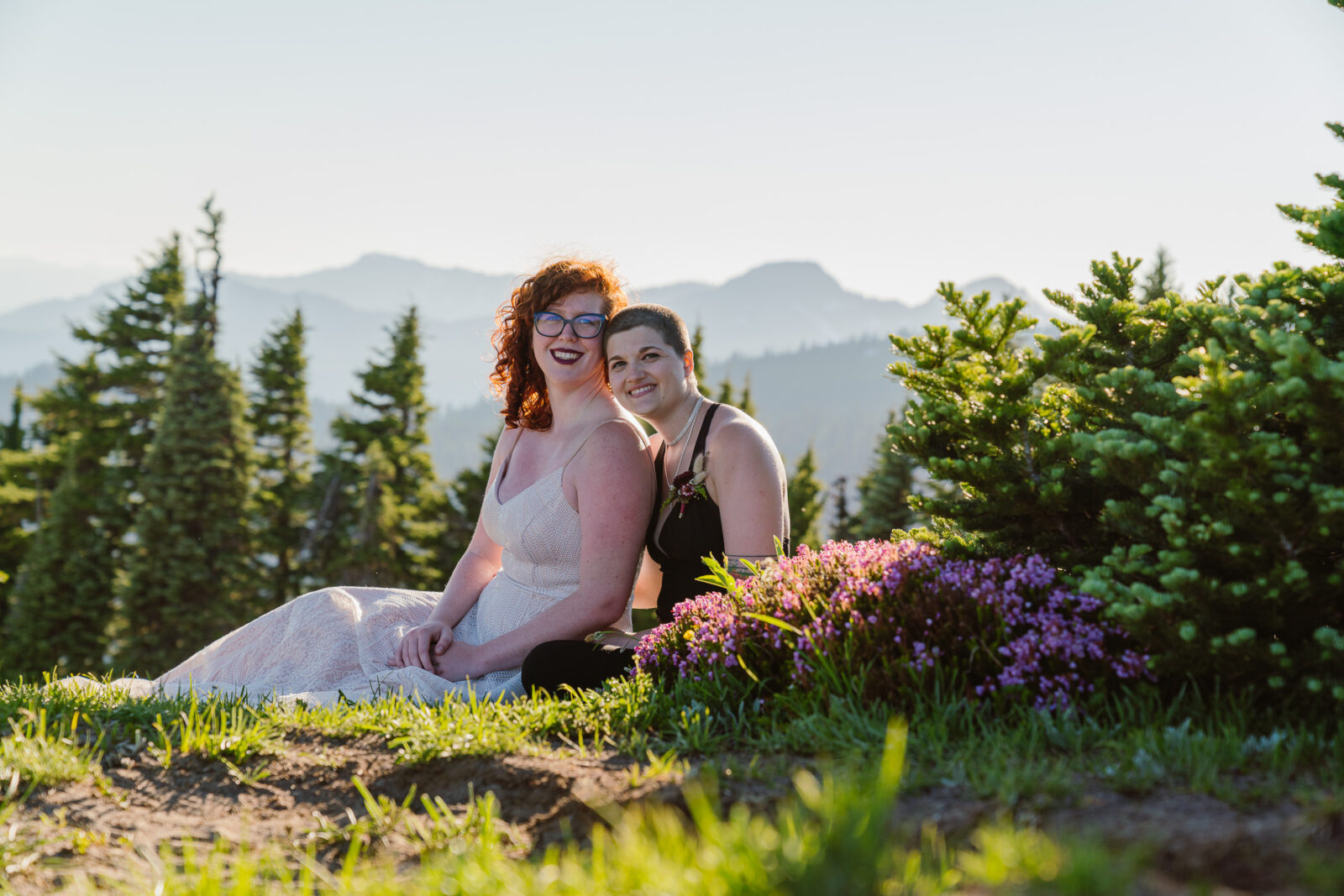 Couple sits on ground behind purple wildflowers at Mount Rainier National Park as the sun sets