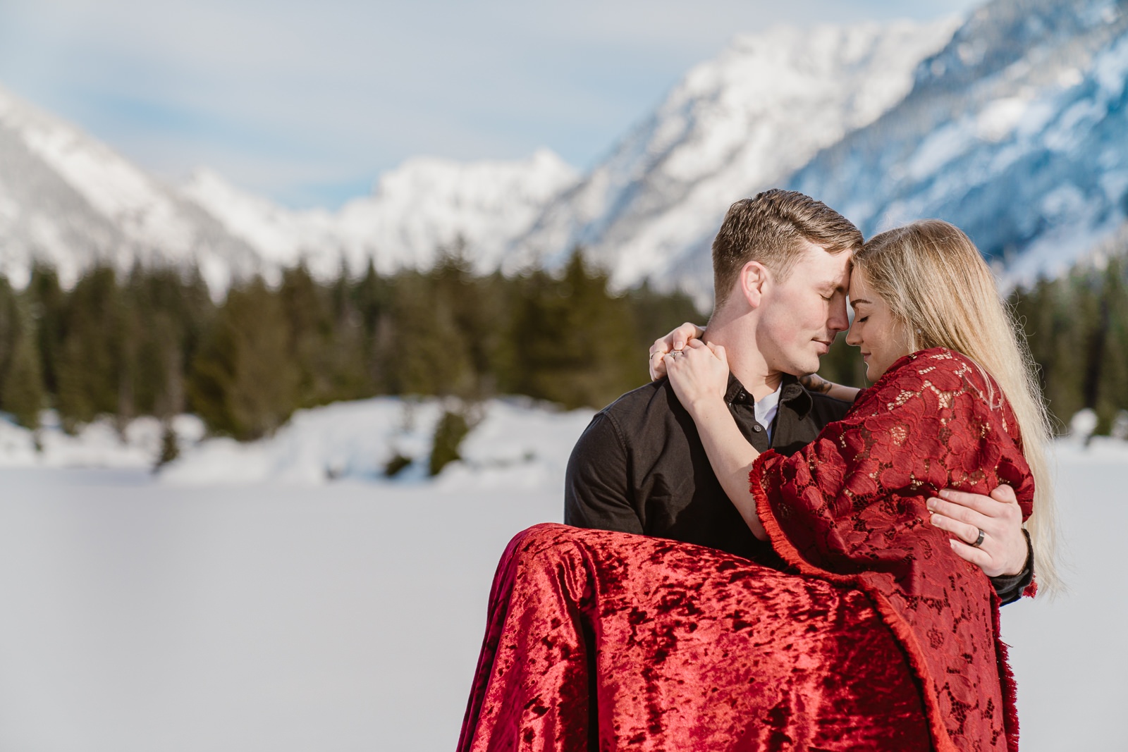 Husband holds wife in his arms at a snow covered alpine lake with mountains in the background